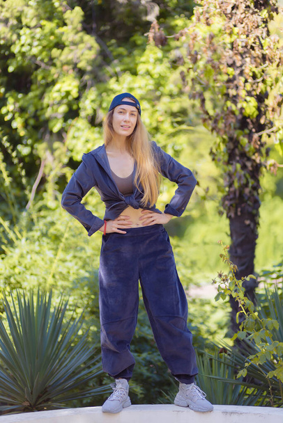 Langhaarige Blondine im Hip-Hop-Stil steht im Park vor dem Hintergrund südländischer Pflanzen - Foto, Bild