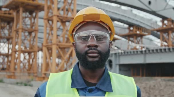 Tracking portrait of African-American male construction site worker in safety vest, hard hat and goggles looking at camera outside. Unfinished building with scaffolding in background - Footage, Video