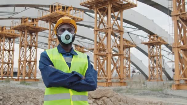 Portret Afroamerykanina męskiego budowlańca w kamizelce bezpieczeństwa, twardej czapce i masce przeciwpyłowej do kamery przed niedokończonym budynkiem z rusztowaniami - Materiał filmowy, wideo