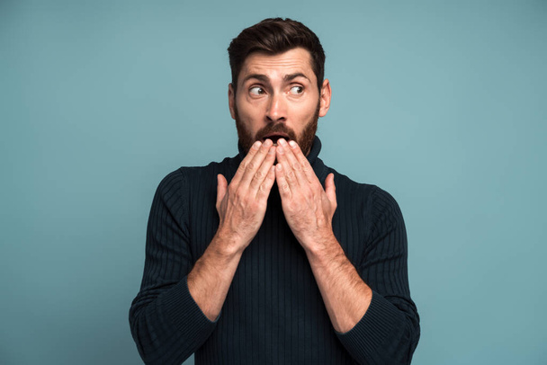 Wow. Man covering mouth with hand and looking away with big eyes, scared surprised expression, shocked by sudden news. Indoor studio shot isolated on blue background  - Photo, Image