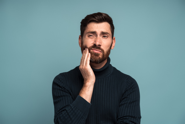 Dental problems. Portrait of unhealthy man pressing sore cheek, suffering acute toothache, periodontal disease, cavities or jaw pain. Indoor studio shot isolated on blue background - Photo, Image
