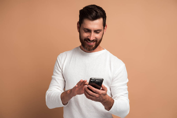 Assertive szakállas férfi figyelmesen nézi kijelző okostelefon ő gazdaság, szörfözés internet, csinál online vásárlás. Fedett stúdió lövés elszigetelt bézs háttér - Fotó, kép