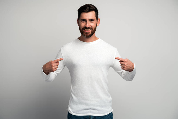This is me. Portrait of conceited man in white shirt pointing at himself and looking with arrogant selfish expression, feeling successful and self-important. Indoor studio shot isolated on white background  - Photo, Image