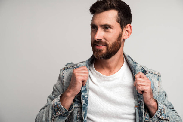 Portrait of bearded handsome man in casual style looking away and smiling while straightening his clothes and posing to the camera. Studio shot on white background  - Photo, Image