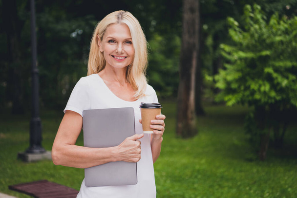 Photo of mature work blond lady hold laptop coffee wear white t-shirt in park alone - Foto, immagini