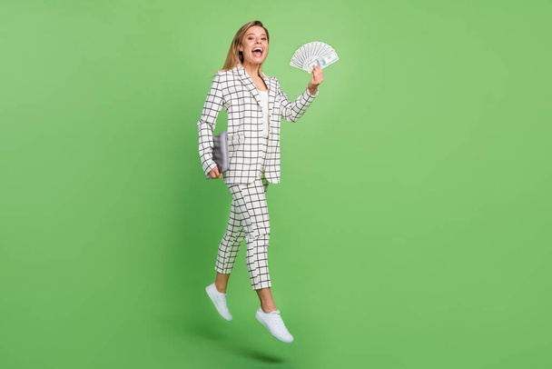 Full body photo of blonde happy woman jump walk hold laptop money wear plaid suit formalwear isolated on green color background - Foto, immagini