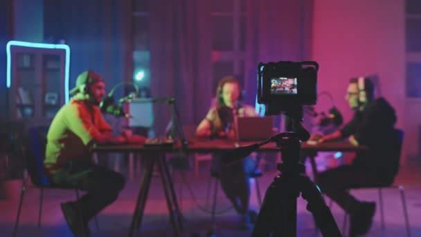 Rastreamento tiro de câmera de vídeo em pé no tripé e filmar três pessoas sentadas à mesa na sala de estar iluminado por luzes de corda de néon e gravação podcast - Filmagem, Vídeo