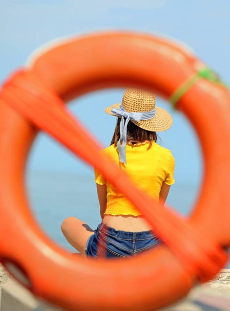 chica joven con sombrero de paja dentro de la boya de la vida intencionalmente fuera de foco por el mar - Foto, Imagen