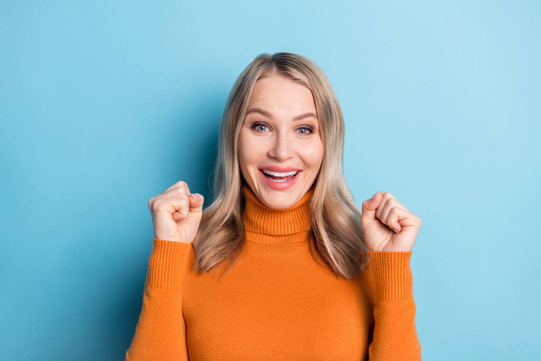 Photo of cute lucky young lady wear orange sweater smiling rising fists isolated blue color background - Fotoğraf, Görsel