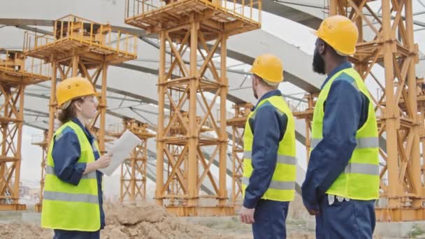 Plan moyen de la superviseure en gilet de sécurité et chapeau rigide tenant le plan directeur et instruisant les travailleurs masculins sur le chantier de construction - Séquence, vidéo