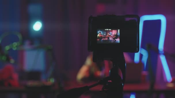 Nahaufnahme mit Fokus der Videokamera, die auf einem Stativ steht und drei junge Leute filmt, die im Wohnzimmer sitzen, beleuchtet von Neonlichtern und Podcast aufnehmen - Filmmaterial, Video
