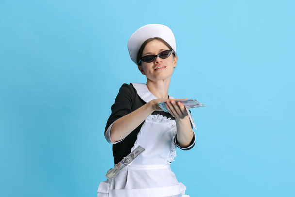 Bossy young woman in chambermaid uniform and stylish sunglasses isolated over blue background - Foto, Imagen
