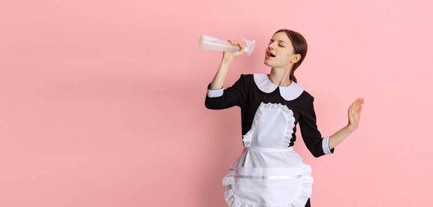 Retrato recortado de jovem bela camareira do hotel em uniforme preto cantando em garrafa de spray de limpeza isolado sobre fundo rosa. Folheto - Foto, Imagem