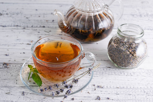 Tea with lavender in a transparent cup on a light wooden table. A glass teapot with tea leaves and a jar with dried lavender flowers in the background - Foto, imagen