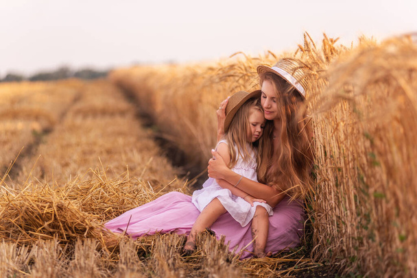 The mother tenderly hugs little daughter, who is sitting on knees by the golden wheat field. Young woman lovingly strokes a girl. The girls sit by the mown rye field. Weekend walks in the countryside - Фото, изображение
