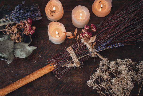 Escoba decorada de bricolaje para la celebración de Samhain. Escoba hecha a mano sobre una mesa de madera oscura con velas blancas encendidas y coloridas flores secas y hierbas en el fondo - Foto, imagen