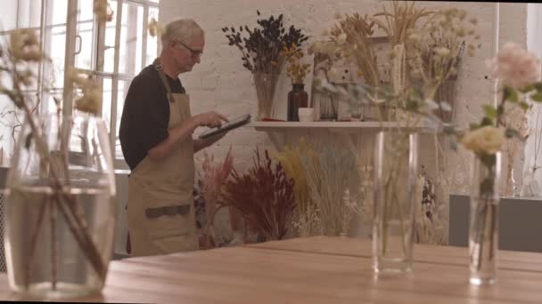 Tiro medio del trabajador de floristería masculino senior en delantal haciendo el inventario de hermosas flores secas en jarrones tomando notas en la tableta digital - Metraje, vídeo