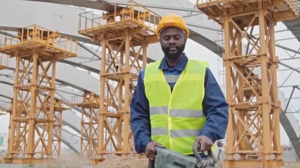 Portret Afroamerykańskiego robotnika budowlanego w kamizelce bezpieczeństwa i twardej czapce z młotem pneumatycznym oraz pozowanie do kamery przed niedokończonym budynkiem z rusztowaniem - Materiał filmowy, wideo