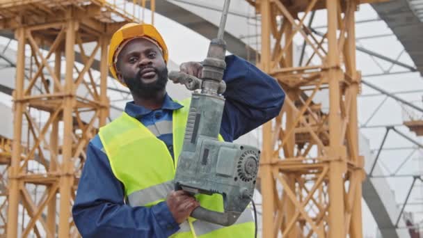 Foto de retrato PAN del trabajador de la construcción afroamericano en chaleco de seguridad y sombrero duro posando con martillo neumático antes de la construcción inacabada con andamios - Metraje, vídeo
