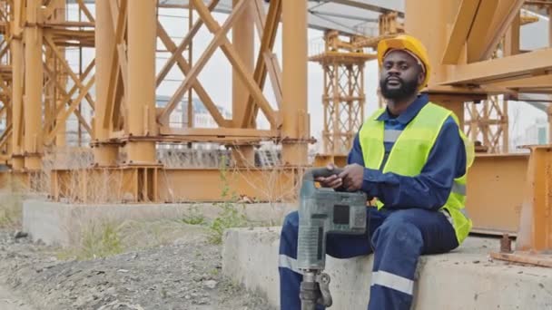Sledování portrétu černocha stavební dělník v bezpečnostní vestě a pevný klobouk drží sbíječku a sedí na betonovém bloku. Odpočívá a dívá se do kamery - Záběry, video