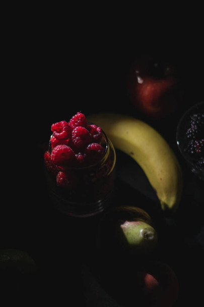 Diferentes frutas juntas sobre fondo negro oscuro, bayas, plátano, manzana, frambuesa, arándano - Foto, Imagen