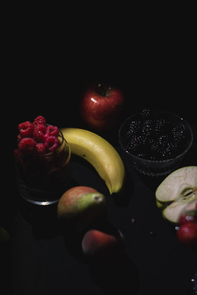 Diferentes frutas juntas sobre fondo negro oscuro, bayas, plátano, manzana, frambuesa, arándano - Foto, Imagen