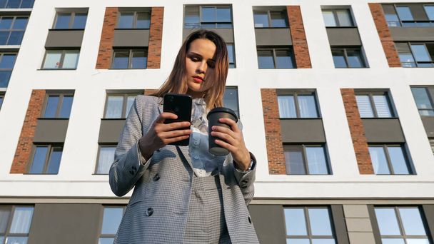 Takım elbiseli bir kadın plastik bardaktan kahve içer ve bir binanın arka planındaki telefona bakar. - Fotoğraf, Görsel