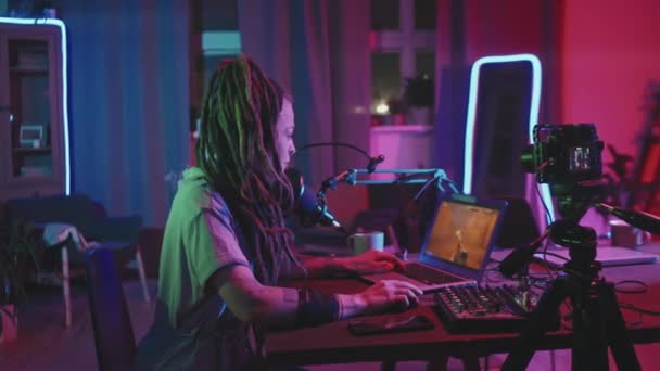 Sledování záběr mladé ženy s dredy sedí u stolu v obývacím pokoji osvětlené struny světla a mluvit do mikrofonu, zatímco hraní a streaming sebe na internetu - Záběry, video