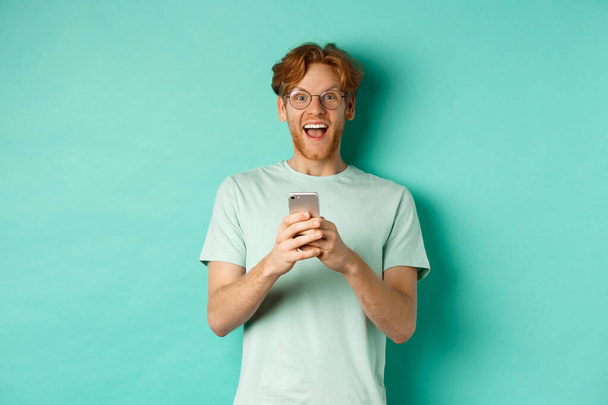 Surpris rousse homme en lunettes regardant étonné de la caméra après avoir lu l'offre promotionnelle sur smartphone, debout sur un fond turquoise - Photo, image