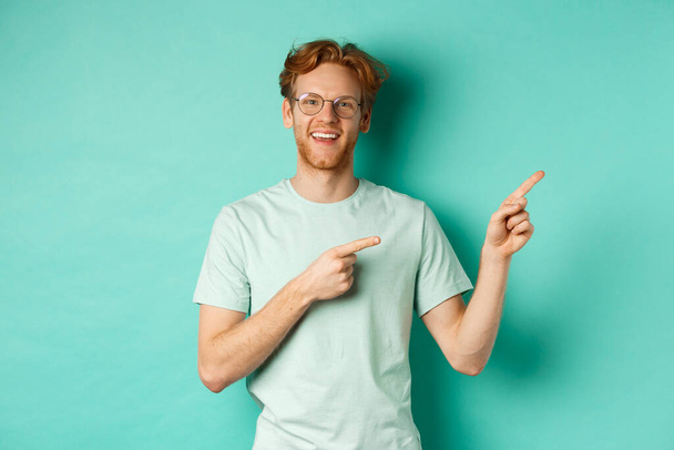 Komea valkoihoinen mies, jolla on inkiväärihiukset, silmälasit ja t-paita, osoittaa sormella oikealle ja hymyilee iloisesti, näyttää mainosta, seisoo turkoosin taustan yläpuolella - Valokuva, kuva