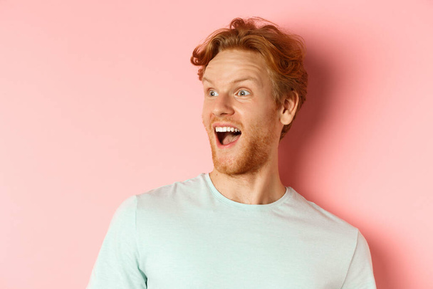 Gros plan de jeune homme heureux et fasciné avec des cheveux roux vérifier promo deal, regardant à gauche avec un sourire étonné, debout sur fond rose - Photo, image