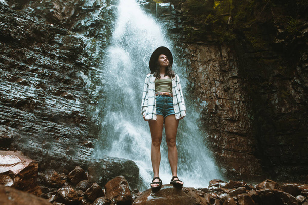 ブラウンハットの女性は滝のハイキング旅行のコンセプトを楽しんでいます - 写真・画像