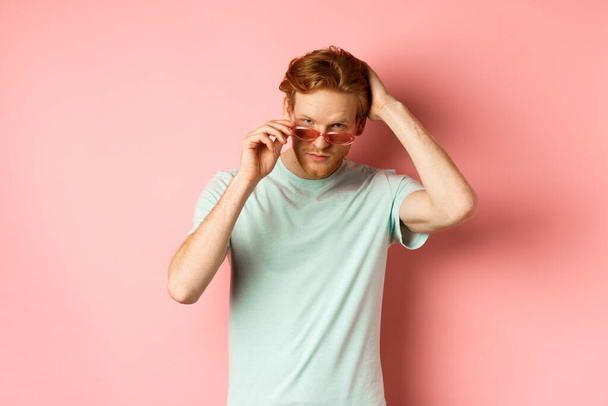 Όμορφος νεαρός κοκκινομάλλης με γυαλιά ηλίου, βούρτσισμα των μαλλιών με το χέρι και αναζητούν αυτάρεσκη και αυτοπεποίθηση στην κάμερα, στέκεται πάνω από ροζ φόντο - Φωτογραφία, εικόνα