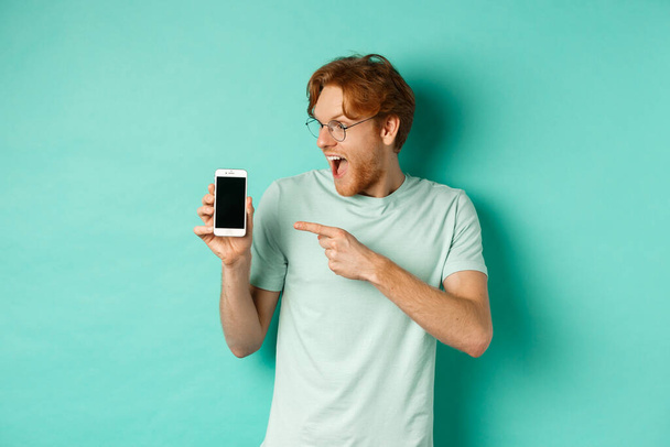 Check this out. Handsome redhead guy in glasses pointing finger at blank smartphone screen, showing online promotion, standing amazed over turquoise background - Foto, immagini