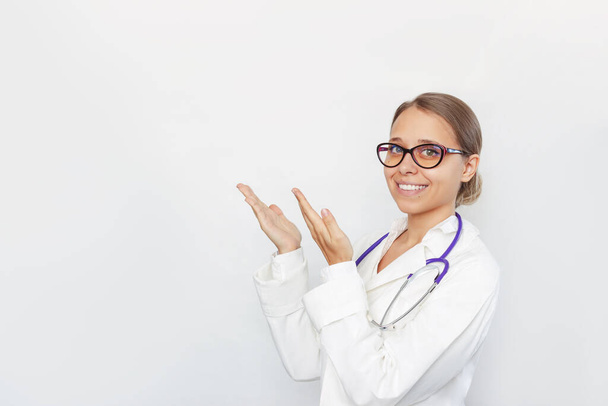 A young blonde female doctor in a white coat and glasses with a stethoscope around her neck indicates an empty copy space for text or design with her hands isolated on a white background - Zdjęcie, obraz
