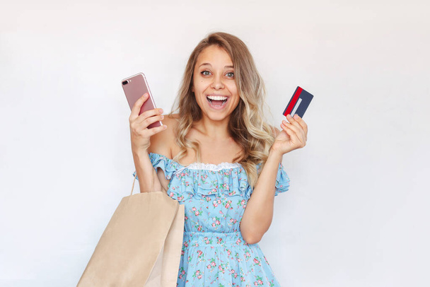 Dalgalı saçlı, gülümseyen genç bir kadın beyaz bir arka planda izole edilmiş internet alımlarını ödemek için elinde plastik bir kredi kartı, cep telefonu ve kağıt eko çanta tutuyor. - Fotoğraf, Görsel