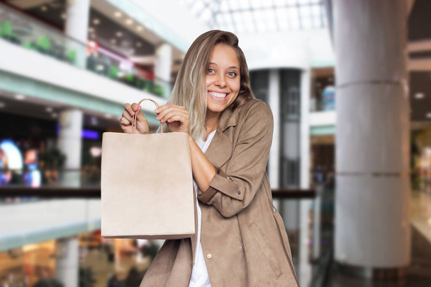 Happy blonde woman with wavy hair with eco bags in her hand on a blurry background of the mall. A smiling girl makes purchases in a shopping center - Fotoğraf, Görsel