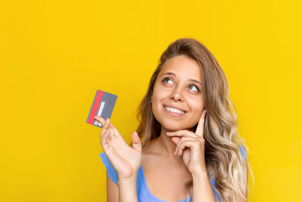 Close-up of a young blonde woman with wavy hair holding a plastic credit card in her hand thinking about how to spend the money looking up at empty copy space isolated on a color yellow background - Photo, Image