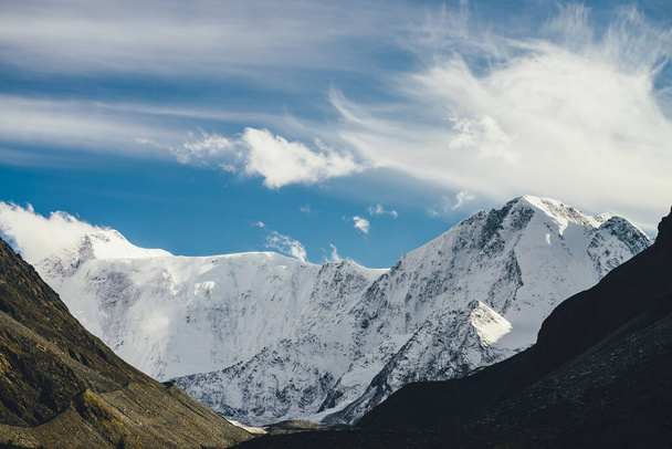 Sfeervol landschap met hoge besneeuwde berg met toppen onder cirruswolken in de lucht. Lage bewolking op grote besneeuwde berg in zonlicht. Zwarte rotsen in gouden zon en wit-sneeuw puntige piek - Foto, afbeelding