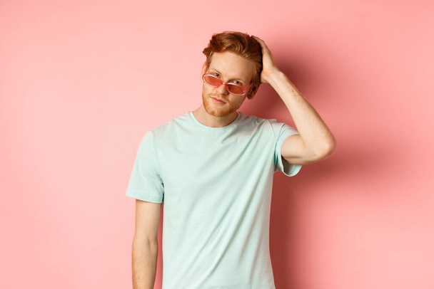 Knappe jonge roodharige man in een zonnebril, borstelen haar met de hand en kijken zelfvoldaan en zelfverzekerd op de camera, staande over roze achtergrond - Foto, afbeelding