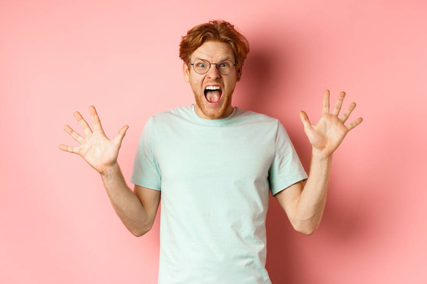 Wütender und bedrängter junger Mann verliert die Nerven, breitet die Hände zur Seite und schreit mit wütendem Gesicht, steht in Brille und T-Shirt vor rosa Hintergrund - Foto, Bild