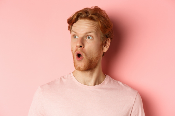 Κοντινό πλάνο του σοκαρισμένου κοκκινομάλλη με γενειάδα, λέγοντας wow, κοιτάζοντας αριστερά με κατάπληκτος πρόσωπο, στέκεται πάνω από ροζ φόντο - Φωτογραφία, εικόνα