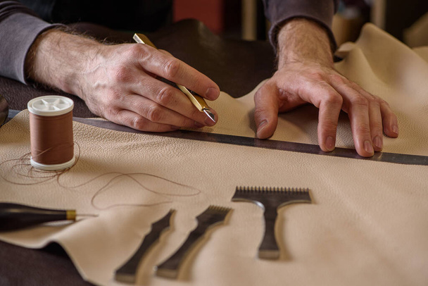 Die männlichen Hände des Schuhmachers halten ein handwerkliches Messer in der Hand, das Leder misst, bevor es in der Werkstatt geschnitten wird. Nahaufnahme - Foto, Bild