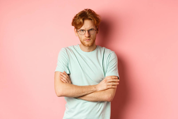 Wütender Mann mit Brille, rotes Haar runzelt die Stirn, verschränkte Arme auf der Brust in defensiver Pose, schmollt dich an, steht über rosa Hintergrund - Foto, Bild
