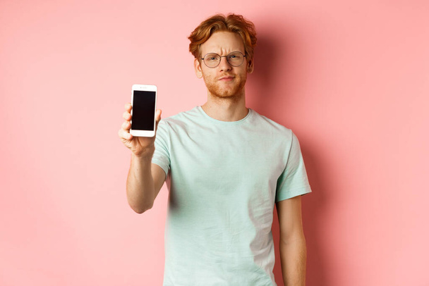 Disappointed male model frowning, showing smartphone screen, standing over pink background - Фото, зображення