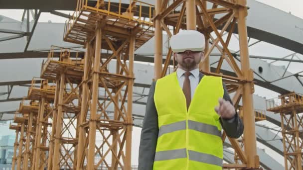 Alacsony szögű lövés férfi helyszíni ellenőr öltöny és biztonsági mellény segítségével virtuális valóság headset előtt befejezetlen épület állványzat - Felvétel, videó