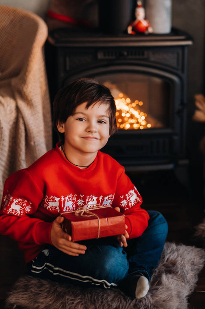 portrait of a smiling boy with a gift in his hands near the fireplace at home. - Photo, image