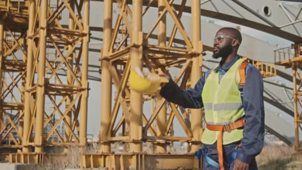 Slowmo mittlere Aufnahme eines afroamerikanischen männlichen Bauarbeiters in Warnweste, Schutzbrille und Gürtel, der sich vor dem Rohbau mit Gerüsten einen harten Hut aufsetzt - Filmmaterial, Video
