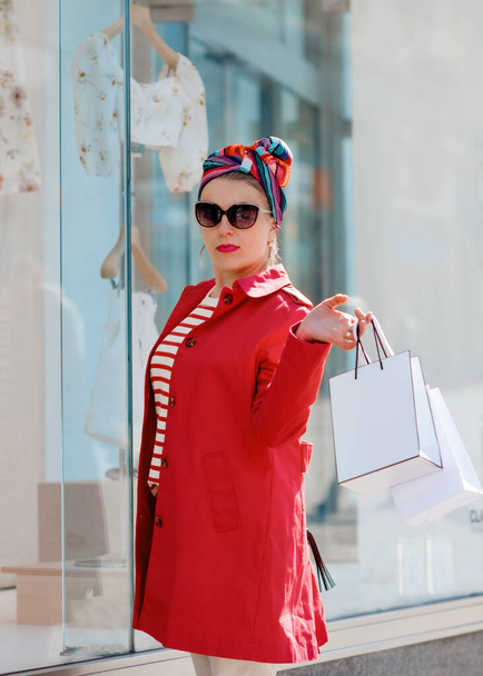 młoda kobieta w czerwonym okopie, okularach przeciwsłonecznych i chustce na głowie robi zakupy i spogląda w witrynę sklepową - Zdjęcie, obraz