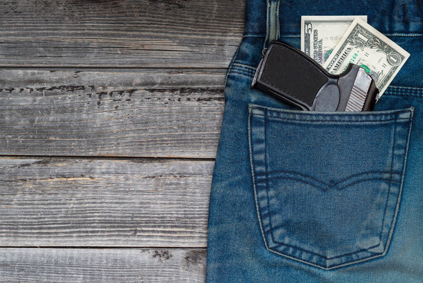 Una pistola policial negra con billetes de dólar en el bolsillo trasero de jeans sobre un fondo de mesa de madera. - Foto, imagen